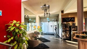 Eine Hotelhalle mit roter Wand, einer Rezeption, einer Sitzecke mit Stühlen, einer großen Topfpflanze und einer Bar mit Flaschen.