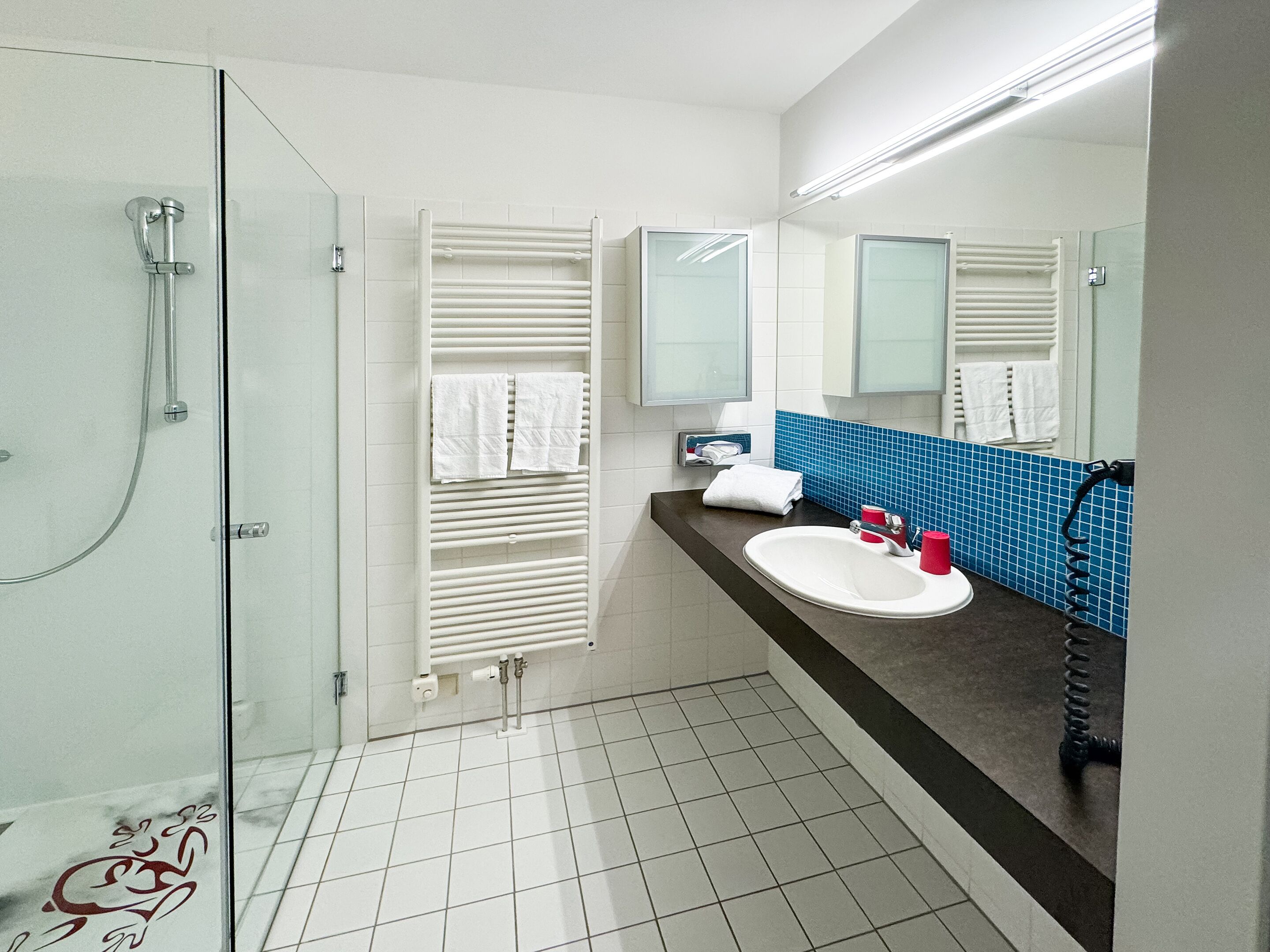 Un baño moderno con cabina de ducha de vidrio, un lavamanos blanco y una taza roja, además de un salpicadero de azulejos azules.