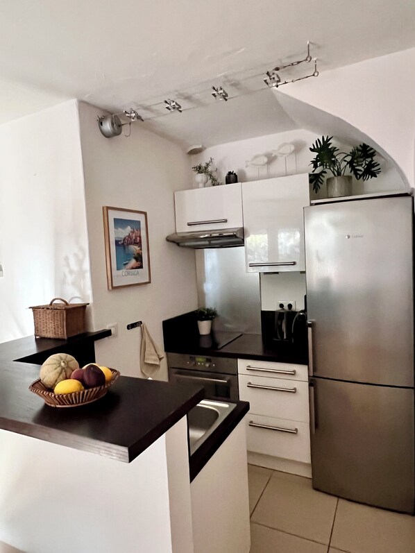 Una cocina compacta con estufa integrada, horno y refrigerador de acero inoxidable. Hay una pequeña isla con un frutero y una cesta.