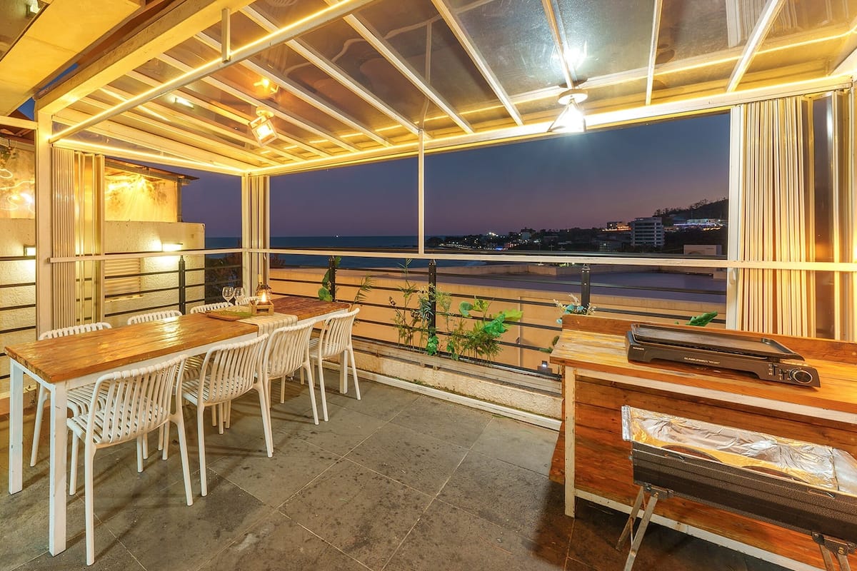 An outdoor dining area with a wooden table and white chairs, a grill, and a view of the city at dusk.