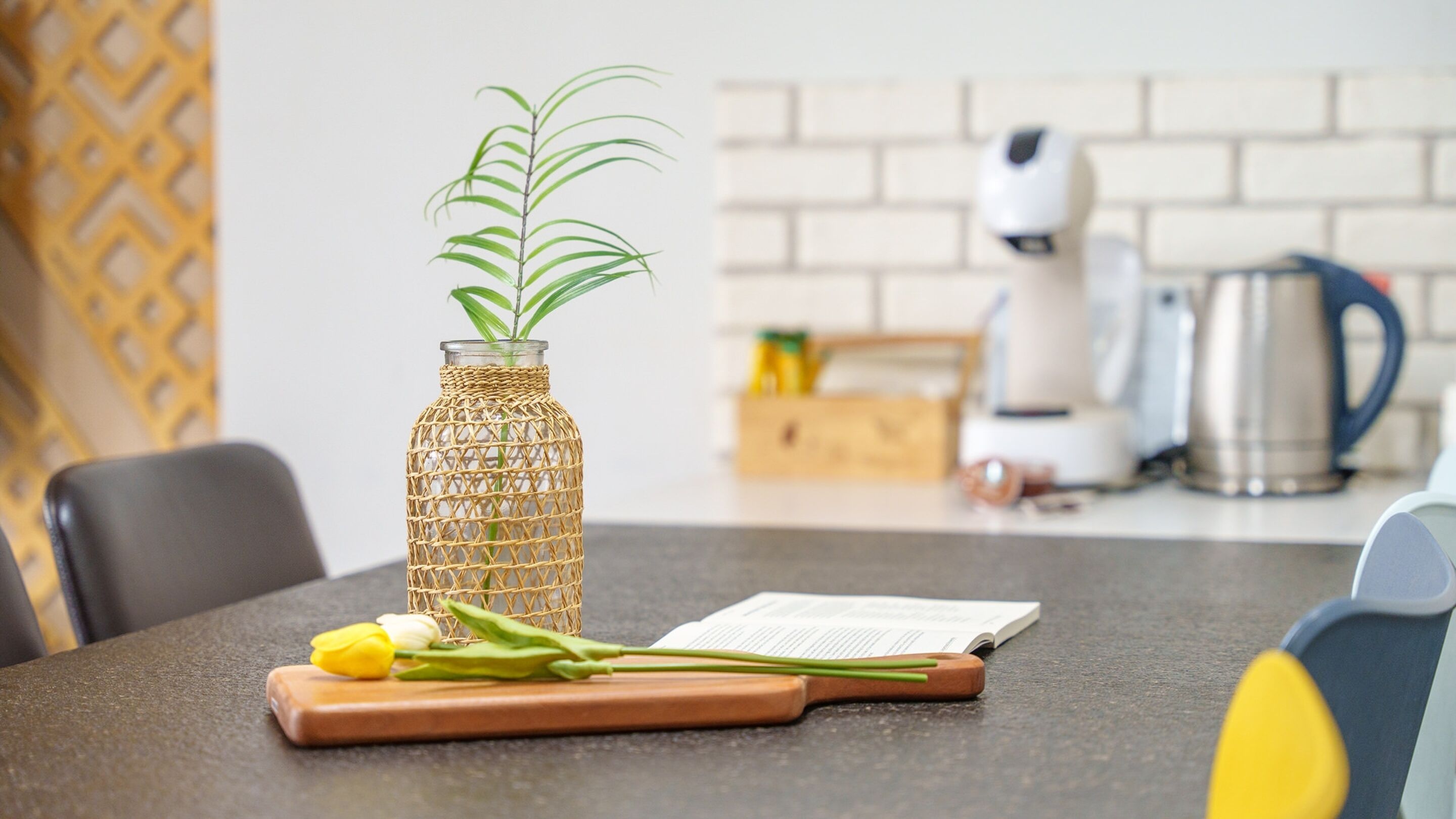 A kitchen counter with a glass bottle containing a plant, a cutting board with a knife, and a book.