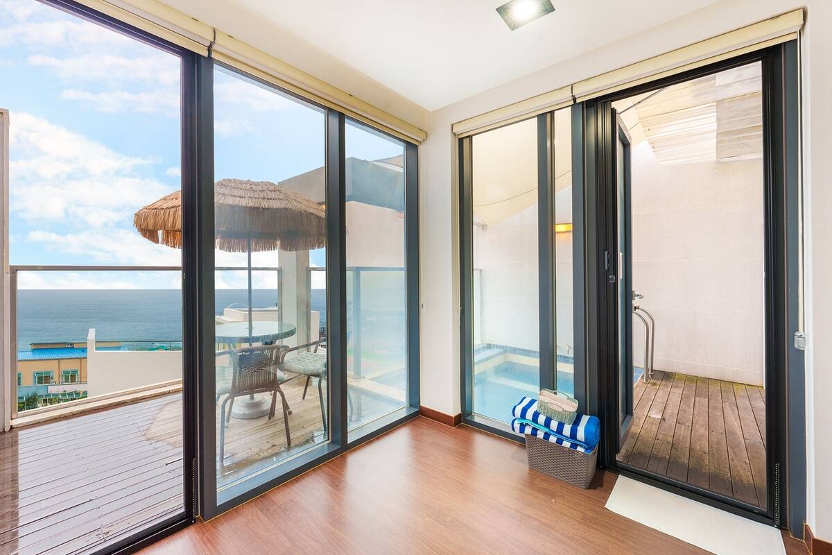 A balcony with a wooden floor, a glass door leading to a pool area, and a view of the ocean.
