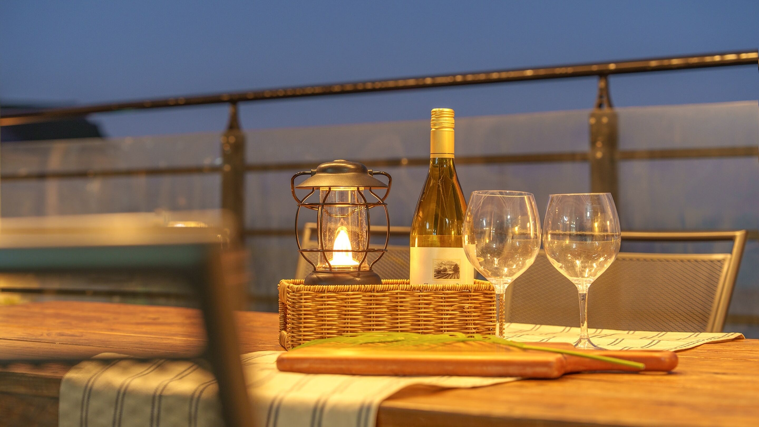 A table with a bottle of wine, two glasses, a candle, and a wicker basket on a balcony with a railing.