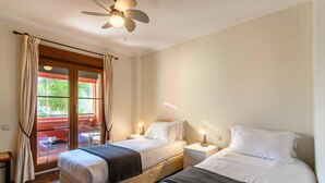 A hotel room with two beds, a ceiling fan, a window with curtains, and a view of greenery outside. - El Rincon del Golf III - Isla Canela Golf GRHIII6P6 (Isla Canela)