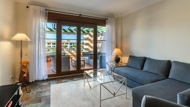 A modern living room with a sofa, a glass coffee table, and a lamp. There is a sliding glass door leading to an outdoor area with a view of buildings and greenery. - El Rincon del Golf III - Isla Canela Golf GRHIII6P6 (Isla Canela)