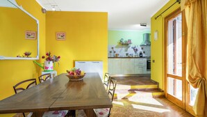 Zona pranzo con tavolo in legno, sedie e un cesto di frutta. Sullo sfondo, una cucina con fornello e pensili.