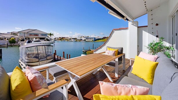 A balcony with a wooden table and chairs, a sofa with cushions, and a view of a marina with boats and houses.