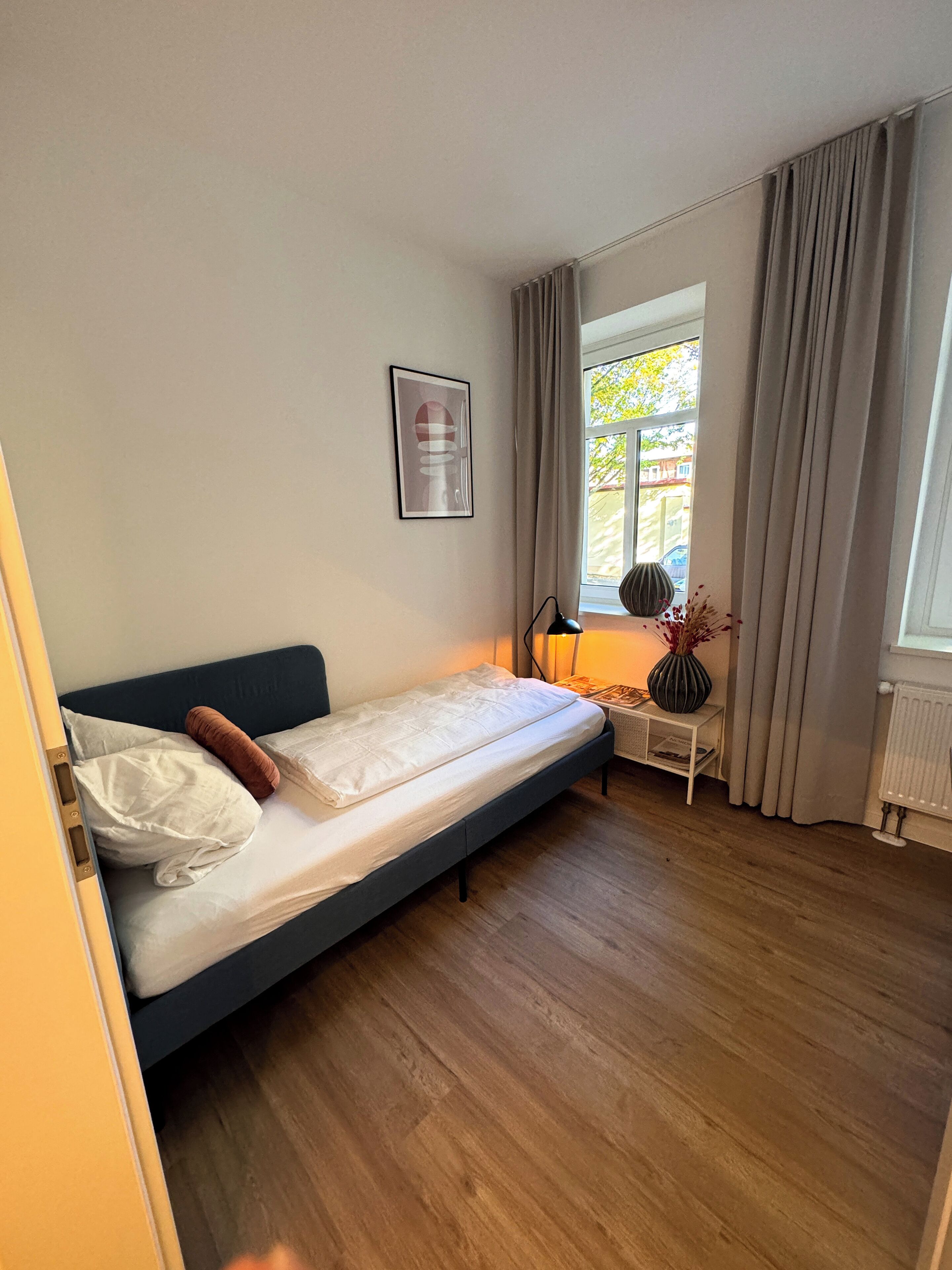 Ein Einzelbett mit Kopfteil, ein Nachttisch mit Lampe, ein gerahmtes Bild an der Wand und ein Fenster mit Vorhängen.