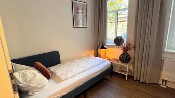 Ein Einzelbett mit Kopfteil, ein Nachttisch mit Lampe, ein gerahmtes Bild an der Wand und ein Fenster mit Vorhängen.