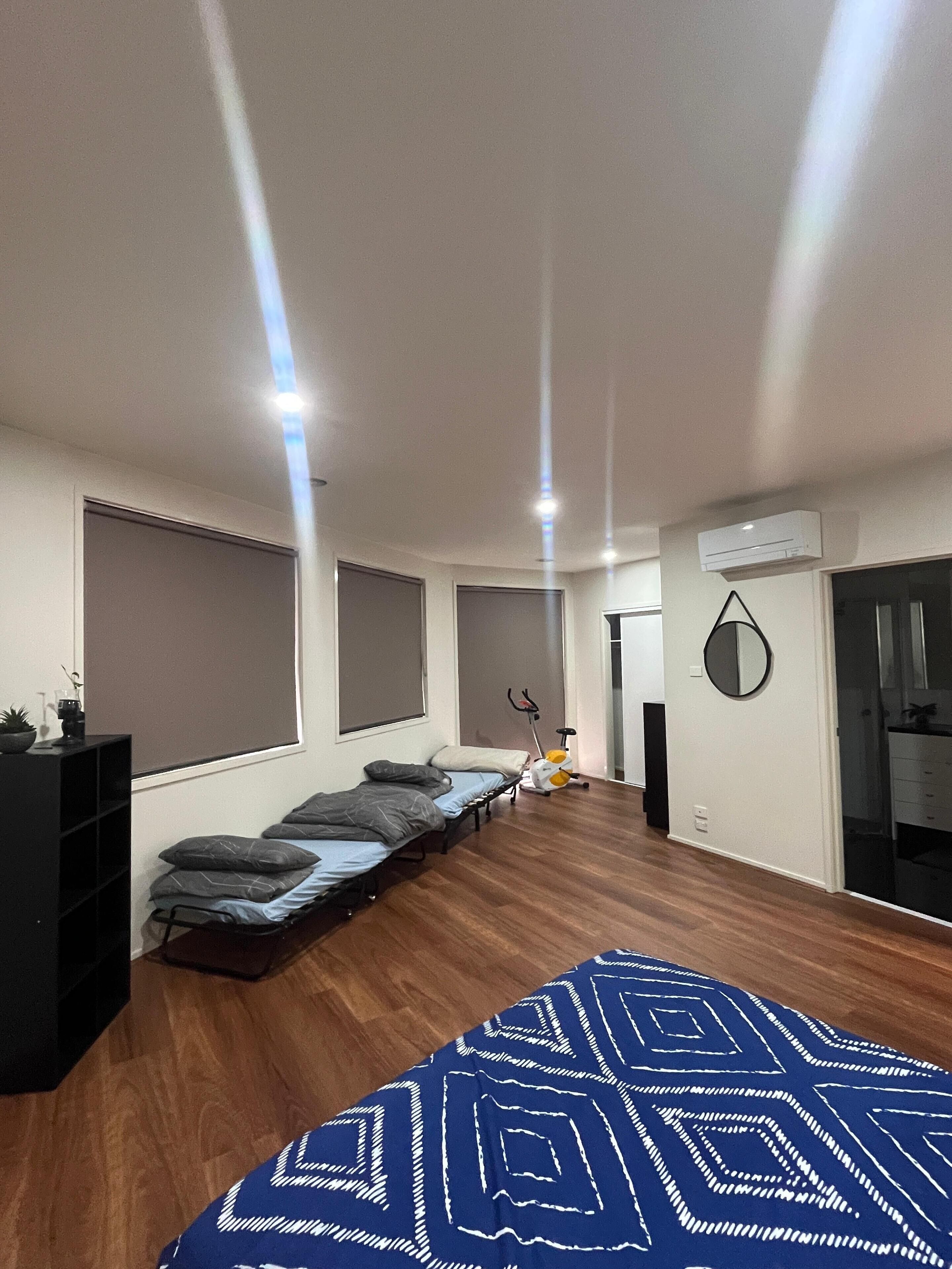 A modern bedroom with a bed, a small desk, a chair, a standing fan, and a window with blinds.