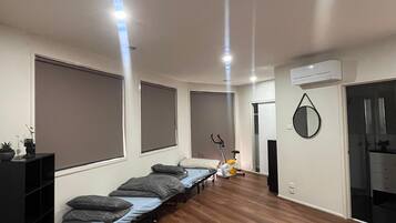 A modern bedroom with a bed, a small desk, a chair, a standing fan, and a window with blinds.