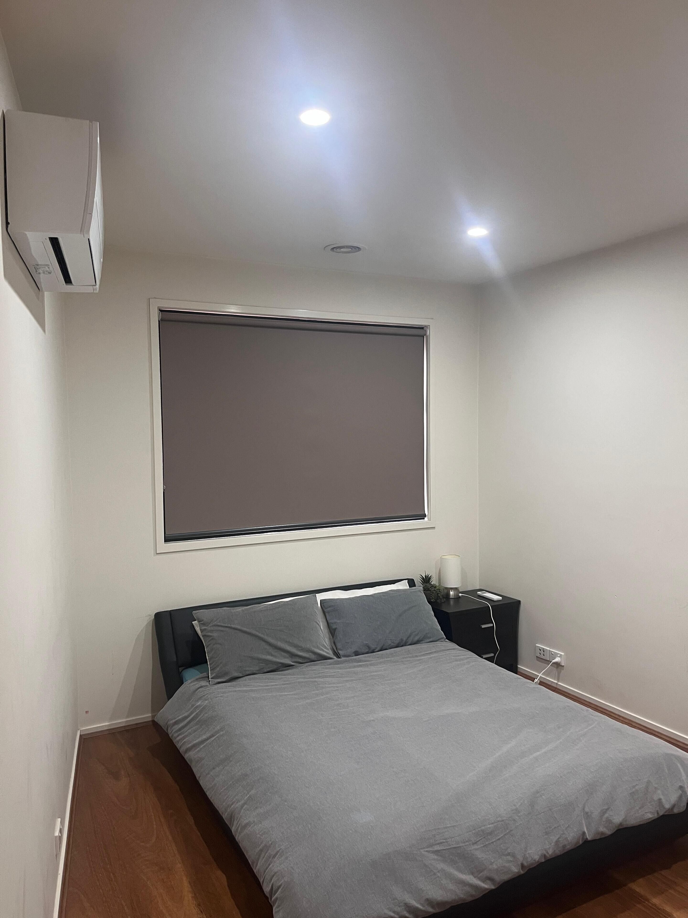 A bedroom with a bed, a bedside table, a nightstand, a wall-mounted air conditioner, and a window with a blind.