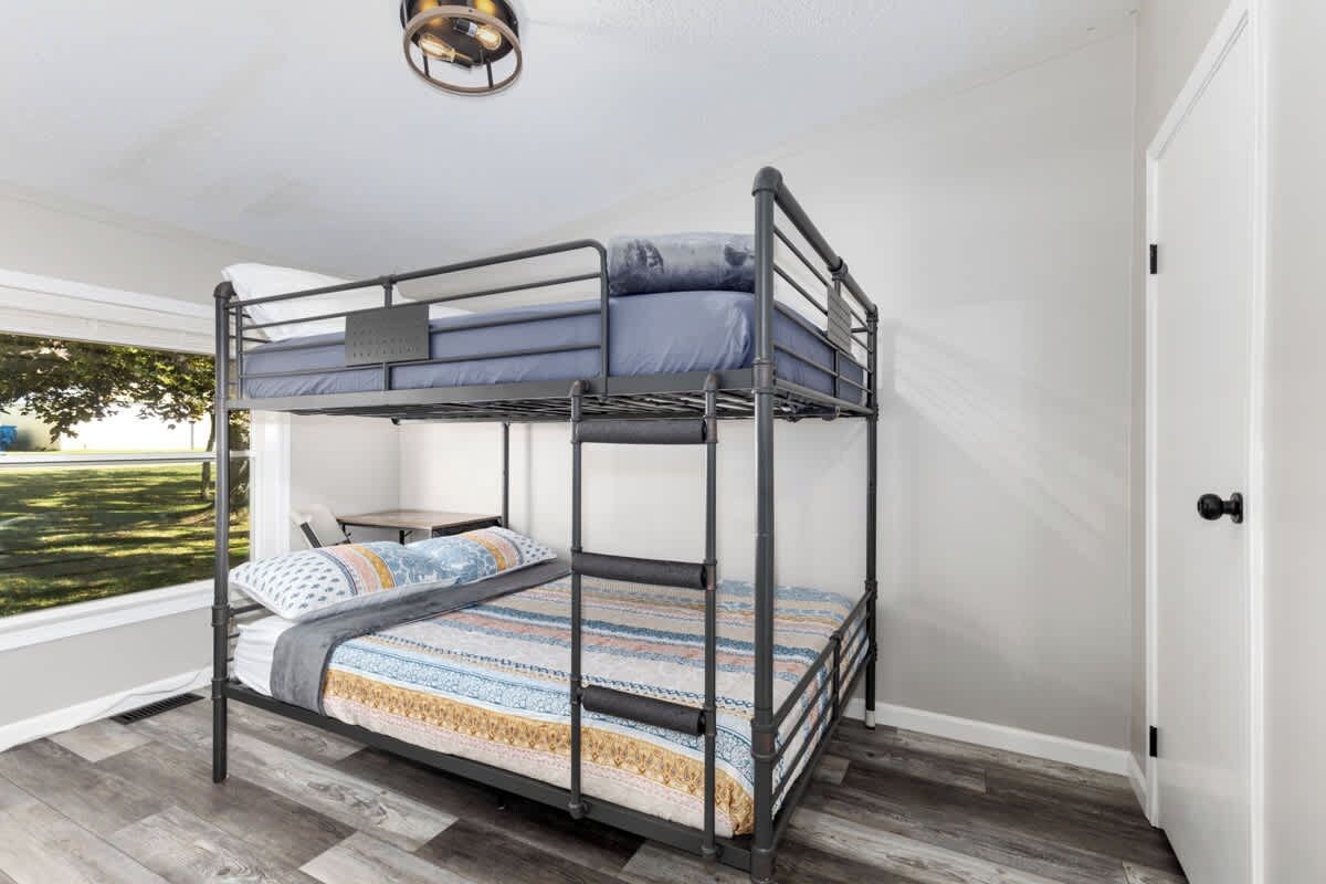 A bunk bed with a patterned bedspread, a small table, and a view of greenery outside.