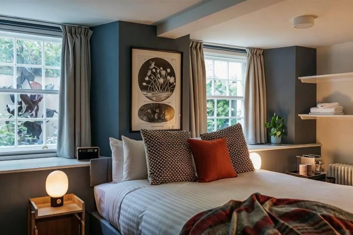 A bedroom with a bed, a bedside table with a lamp, a window with curtains, and a wall-mounted shelf with a plant.