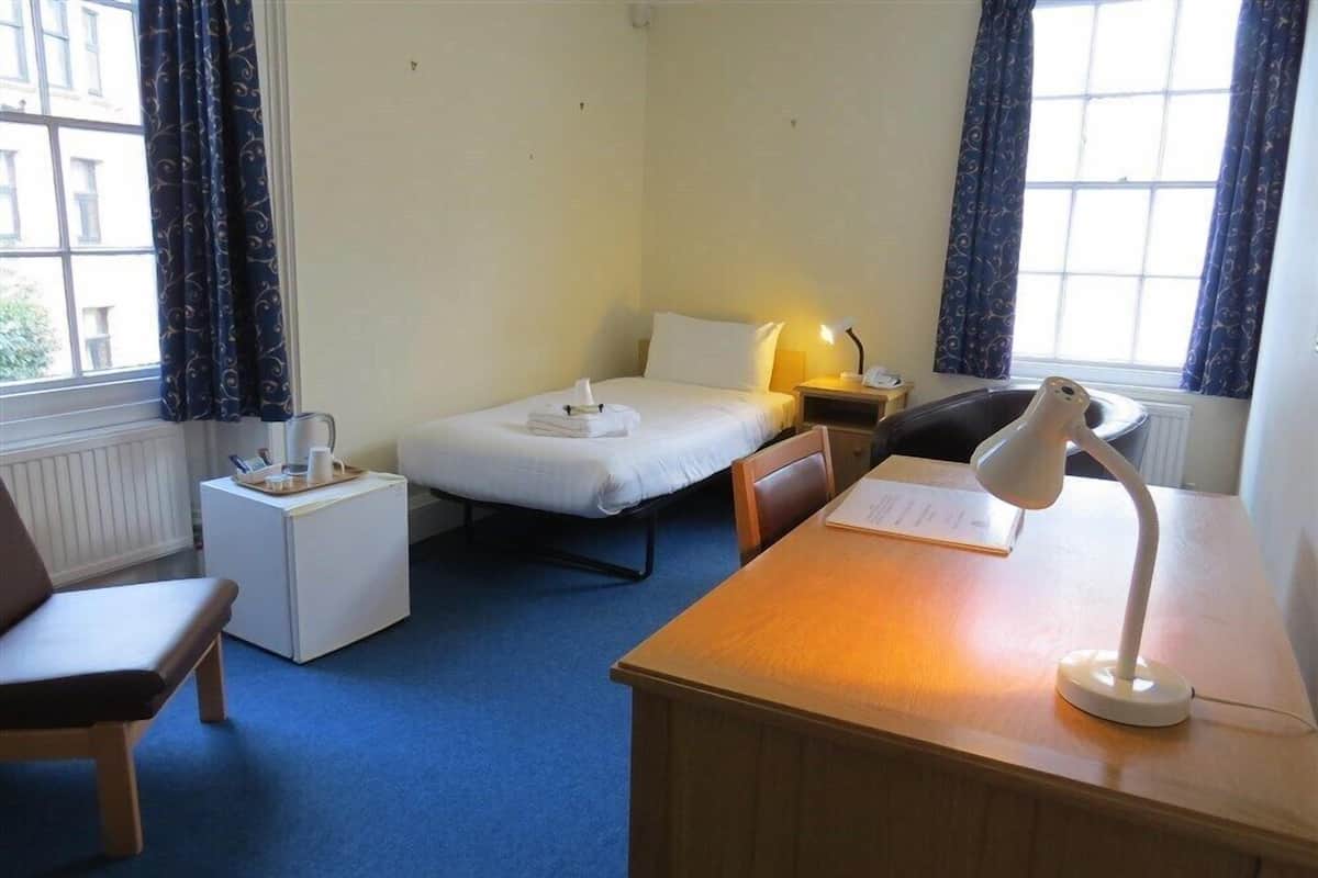 A hotel room with a single bed, a desk with a lamp, a chair, and a small table with a tray.