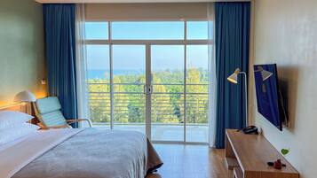 Habitación de hotel con una cama grande, un escritorio, una silla, un televisor y un balcón con vistas a árboles y al mar.