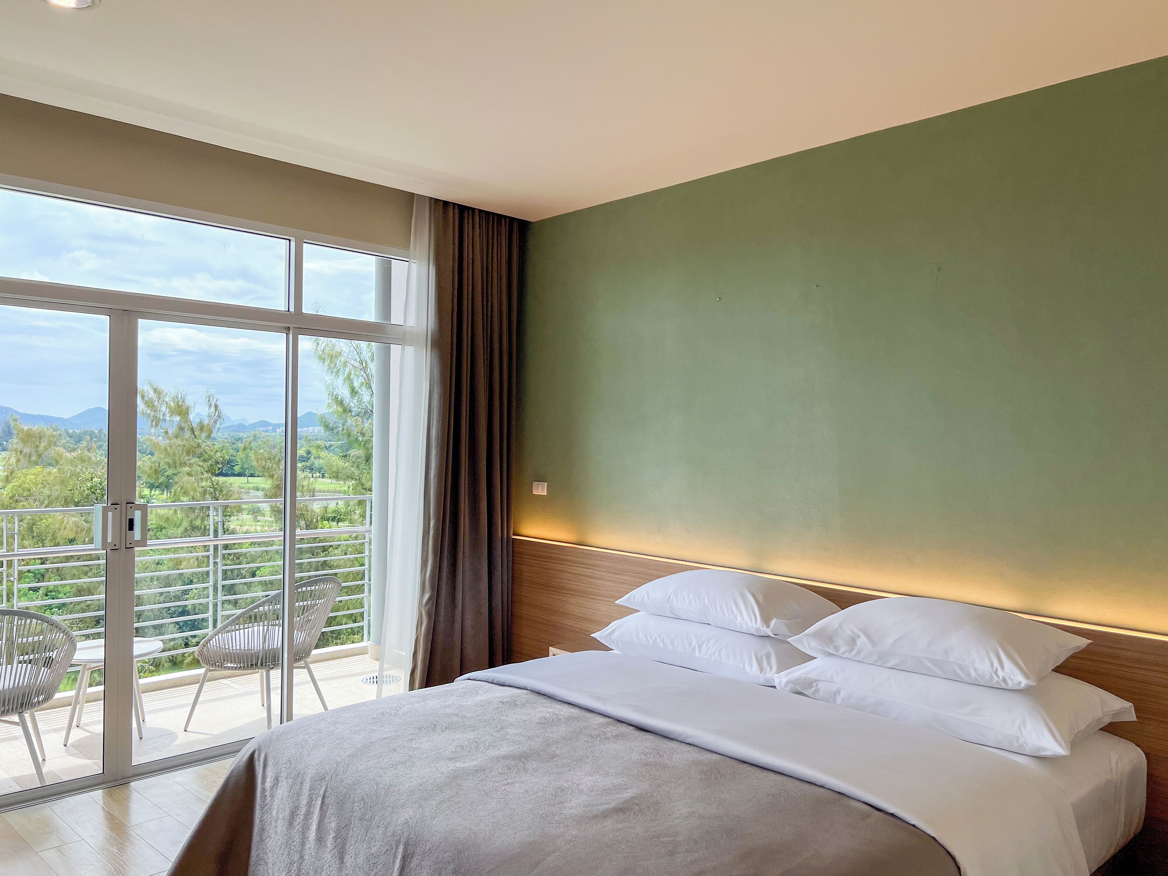 A hotel room with a bed, a view of greenery and mountains through large windows, and a balcony with chairs.