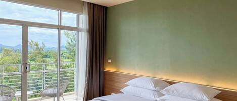 A hotel room with a bed, a view of greenery and mountains through large windows, and a balcony with chairs.