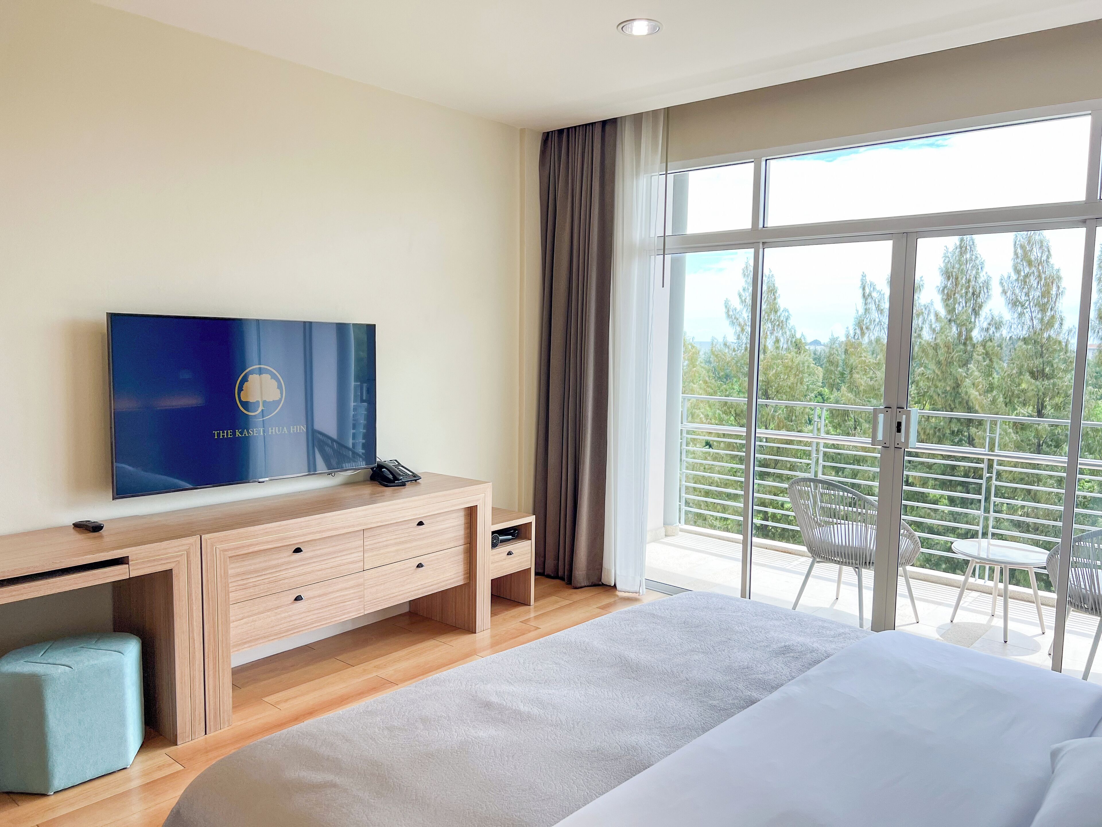 Ett hotellrum med en stor säng, en tv på ett skrivbord i trä, en balkong med utsikt över träd och en skjutdörr i glas.