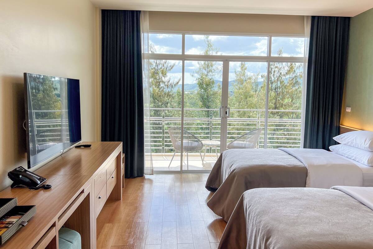 Ein Hotelzimmer mit Bett, Schreibtisch mit Computer, Fernseher und Balkon mit Blick auf Bäume und Berge.