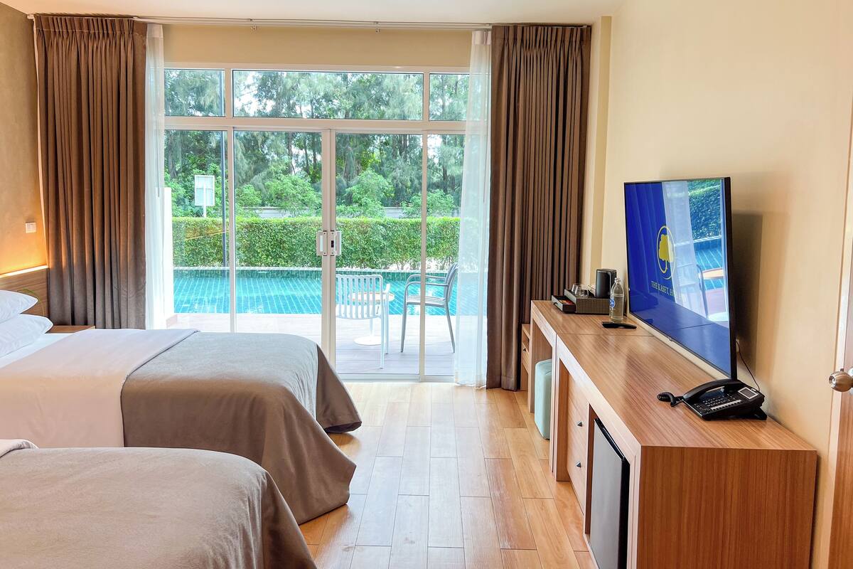 Ein Hotelzimmer mit zwei Betten, einem Fernseher, einem Schreibtisch mit Telefon und Blick auf einen Swimmingpool und Grünflächen.