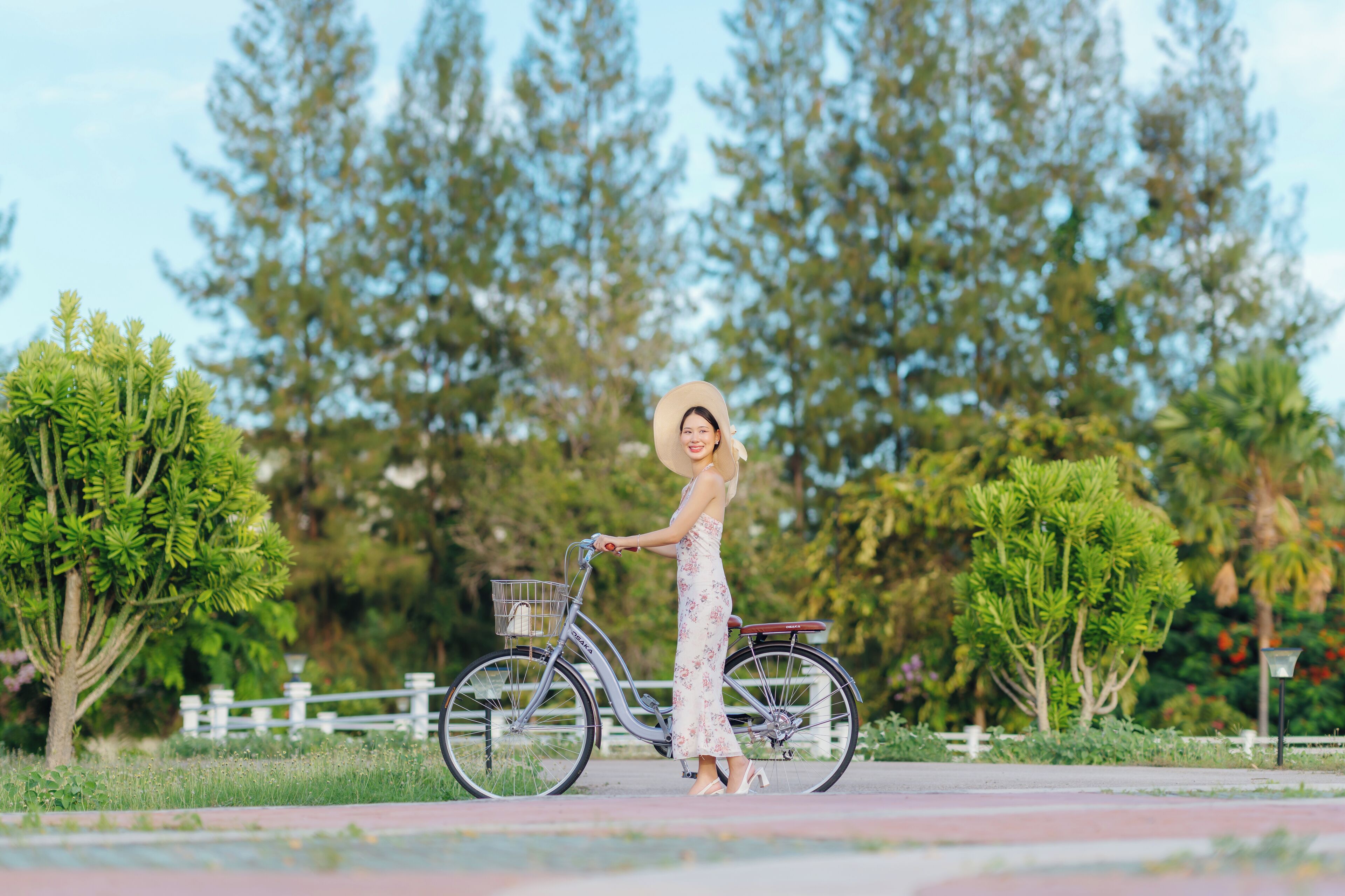 A person standing next to a bicycle with a basket, in a park with trees and a white fence.