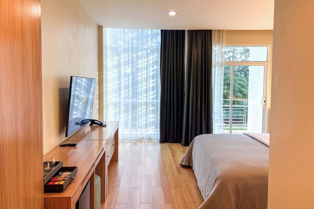Ein Hotelzimmer mit einem Holztisch, einem Flachbildfernseher, einem Bett und einem großen Fenster mit Vorhängen.