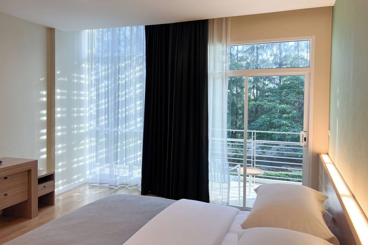 Ein Schlafzimmer mit einem großen Bett, einem Nachttisch, einem Fenster mit leichtem Vorhang und Blick ins Grüne.