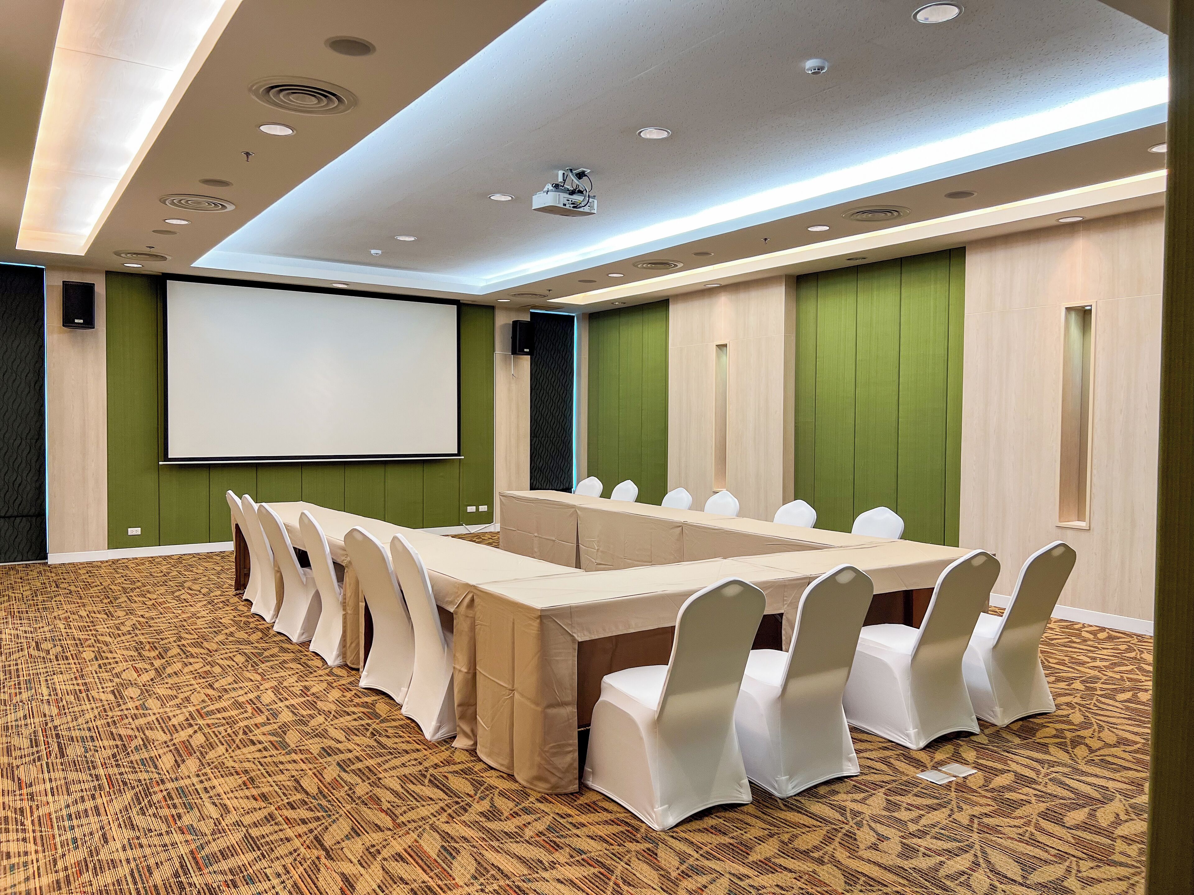 Un salón de conferencias con una larga mesa, sillas blancas, una pantalla de proyector y paredes verdes.