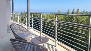 Un balcon avec des fauteuils en osier et une petite table, d’où l’on peut admirer une zone boisée et la mer.