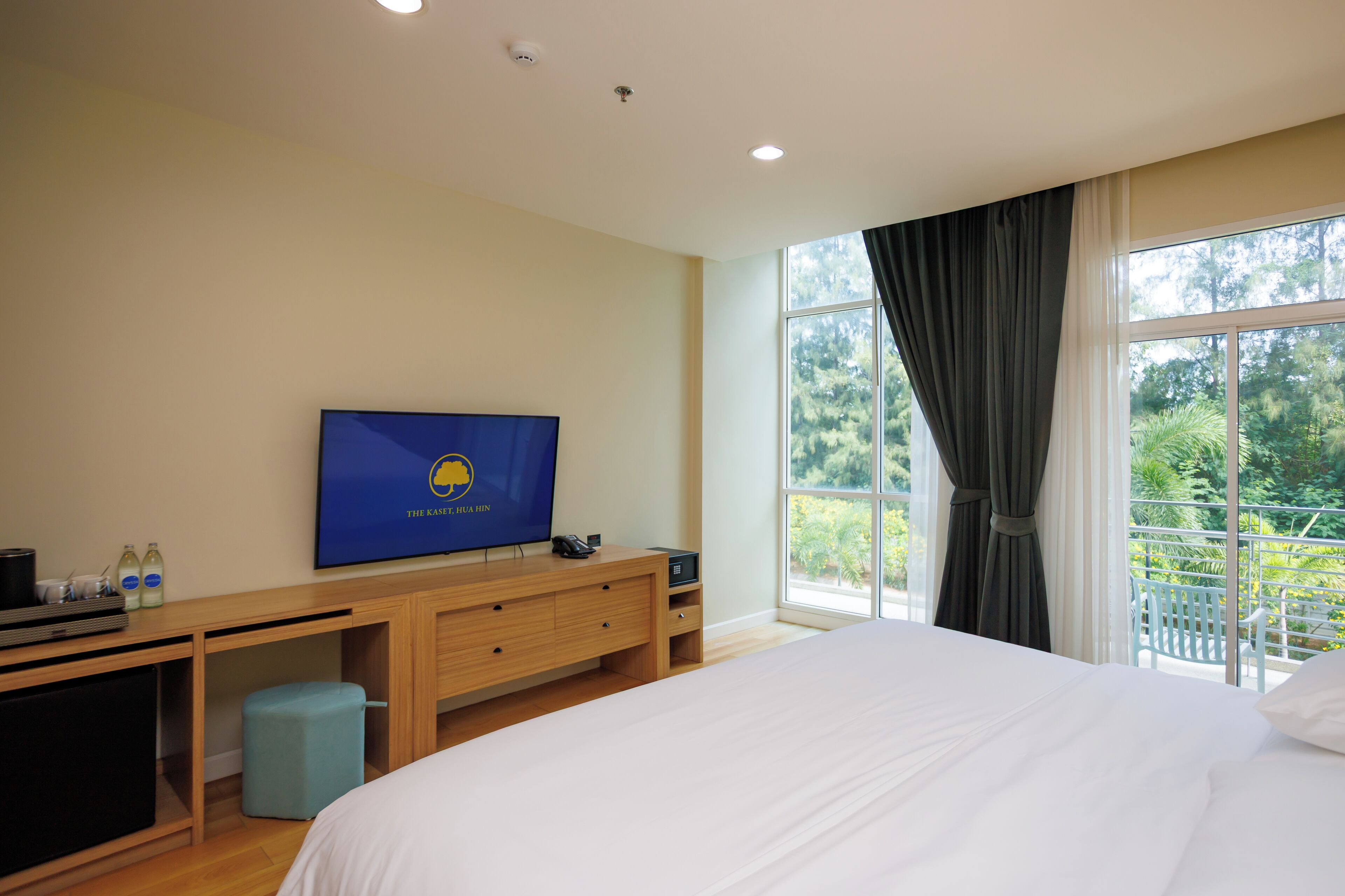 A hotel room with a large bed, a TV on a wooden desk, a small stool, and a balcony with a view of greenery.