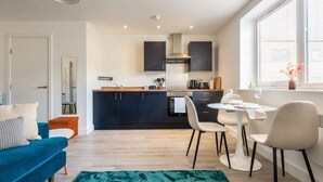 A modern kitchen with dark cabinets, a central island, and a dining area with white chairs and a table. - Bright & Spacious 1 Bedroom Apartment in Bradford (Bradford)