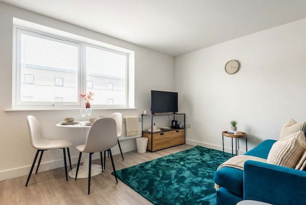 A modern living room with a dining area, a TV on a wooden stand, a clock on the wall, and a blue sofa.