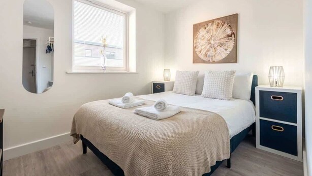 A bedroom with a bed, a window, a wall-mounted mirror, a bedside table, and a wall art piece. - Bright & Spacious 1 Bedroom Apartment in Bradford (Bradford)