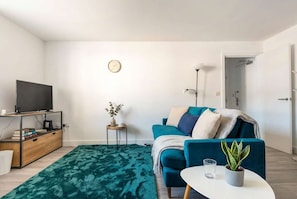 A living room with a teal sofa, a flat-screen TV on a wooden stand, a small side table with a plant, and a green patterned rug.