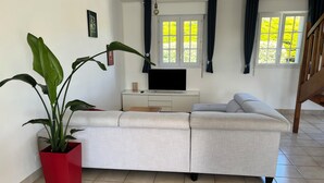 Un salon moderne avec un canapé gris, un pot de fleurs rouge contenant une grande plante, un meuble TV blanc et un escalier.