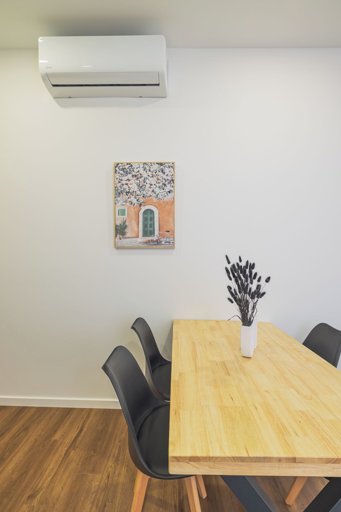 Un comedor con una mesa de madera, sillas negras, un florero blanco con plantas secas y un aire acondicionado de pared.