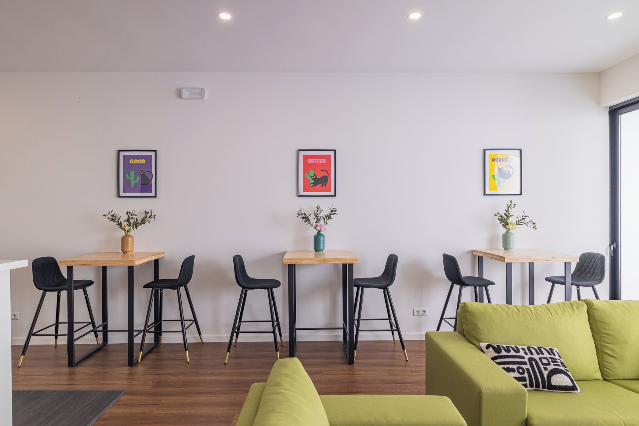 Ein modernes Zimmer mit drei Holztischen, an jedem Tisch jeweils zwei schwarze Stühle. An der Wand hängen drei gerahmte Poster mit unterschiedlichen Motiven. Im Vordergrund steht ein grünes Sofa mit einem gemusterten Kissen.