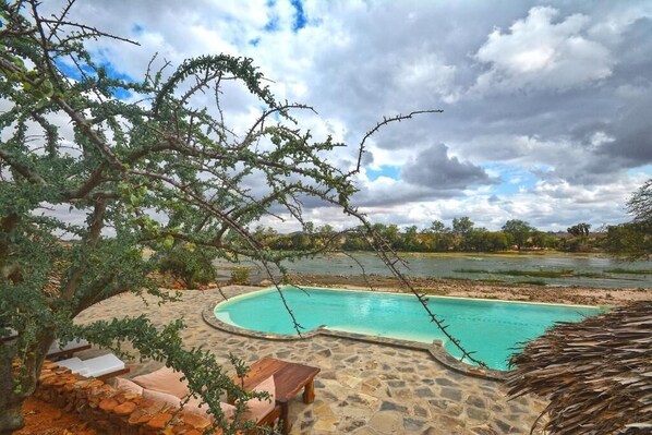 A swimming pool with a stone deck, surrounded by trees and a view of a lake. - Kiboko River Camp (Tsavo East)