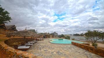 Una piscina de piedra con sillas de descanso y sombrillas de paja, rodeada de muros de piedra y árboles.