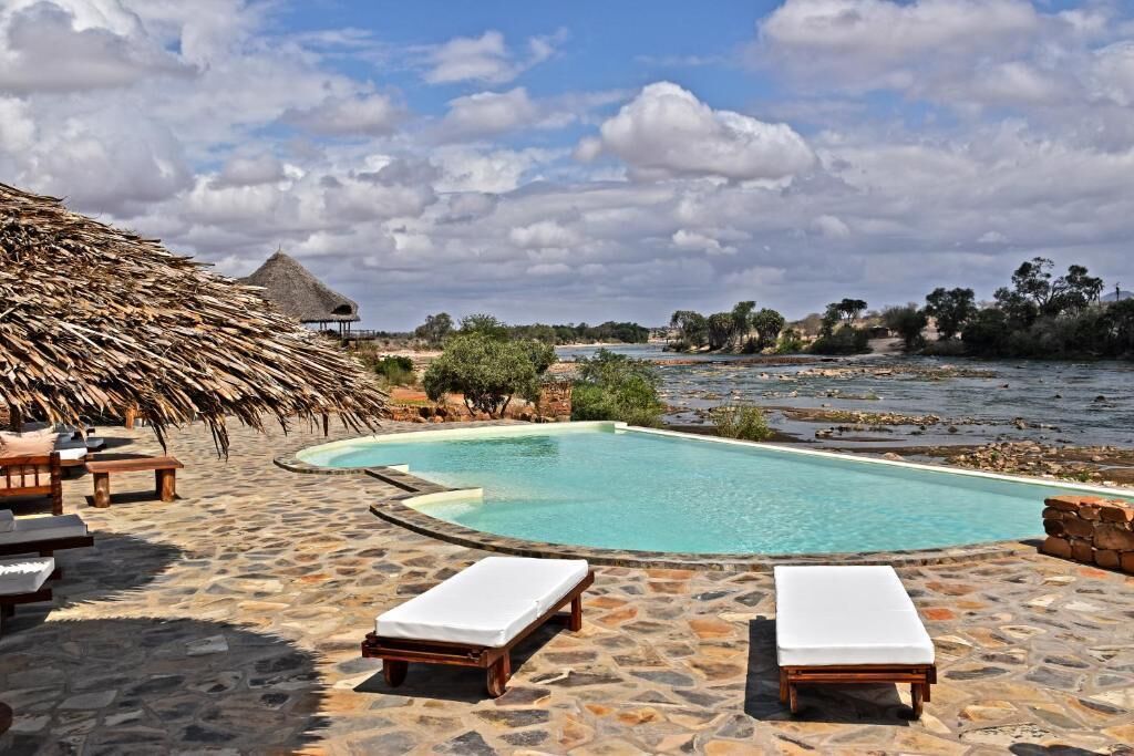 A pool area with lounge chairs, a thatched-roof structure, and a view of the water.