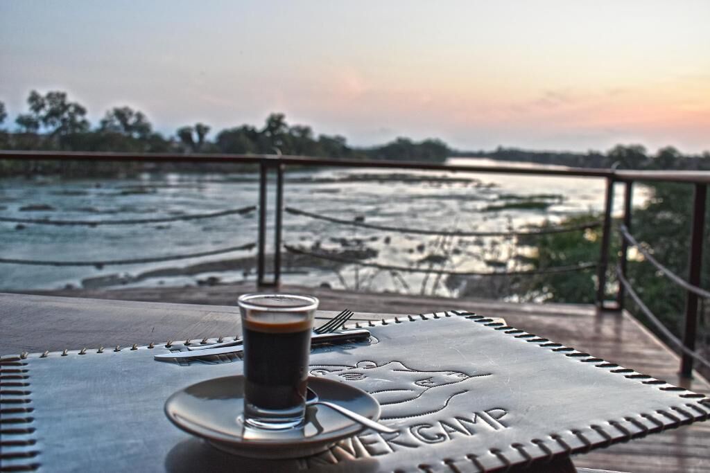 Ein Tisch mit einer Kaffeetasse, einem Löffel und einem Notizbuch auf einer Metallplatte. Im Hintergrund sieht man einen Fluss und Bäume.