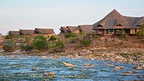 A series of thatched-roof buildings by a river, with rocky terrain and sparse vegetation.