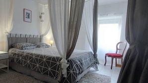 Une chambre à coucher comprenant un lit, une chaise, une table de chevet, une lampe et une fenêtre avec des rideaux.