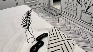A bedroom with a bed, a bedside table with a plant and slippers, a mirror, and a wall with a suitcase and plant decoration.