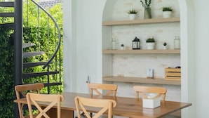 Un coin repas avec une table et des chaises en bois, un ventilateur de plafond, ainsi que des étagères ornées de plantes et d’objets décoratifs.
