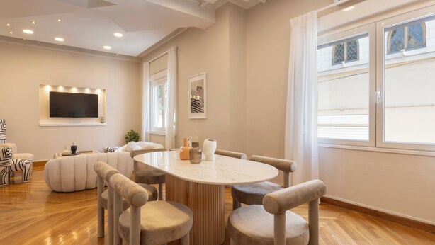 Un salón moderno con una mesa redonda, sillas color beige, un televisor de pantalla plana y un amplio ventanal.