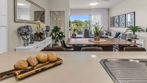 Et moderne spiseområde med et træbord, stole og et fad med brød. Der er et stort spejl på væggen, en loftsvifte og udsigt til grøn bevoksning udenfor.