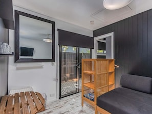 A room with a dark-colored sofa, a wooden bookshelf, a television mounted on the wall, and a sliding glass door leading to an outdoor area. - Welcome to your perfect holiday retreat! (Tweed)