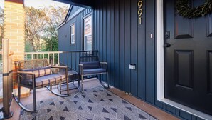 A dark blue exterior with the number 1991, a black door, and a porch with two wicker chairs and a patterned rug. - The Corner House (Huntsville)